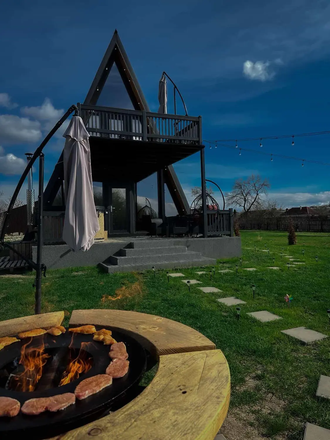 Grătar cu plită în curte, cu fripturi la foc și cabana A-frame în fundal